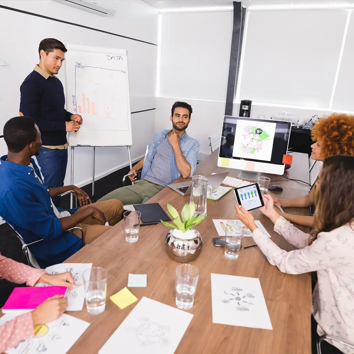 Diverse team of business professionals collaborating in a meeting, with one man presenting data on a whiteboard and a woman holding a tablet.