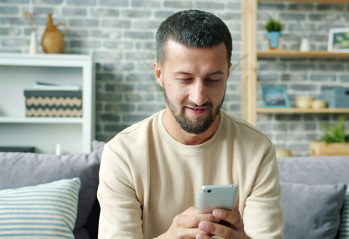 A young man with a beard sitting on a couch, smiling while looking down at and using his smartphone in a modern, brightly lit living room.